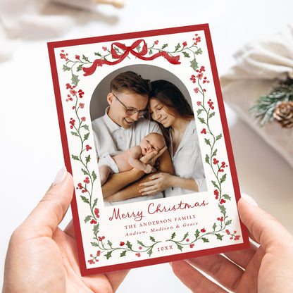 A personalized Christmas family photo card held in two hands. The card features a red border, a red ribbon at the top, and a border of red berries and green holly leaves around a central photo of a family of four.